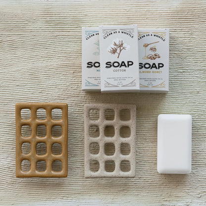 2 colors of ceramic soap dishes arranged on a textured cream surface with bars of soap