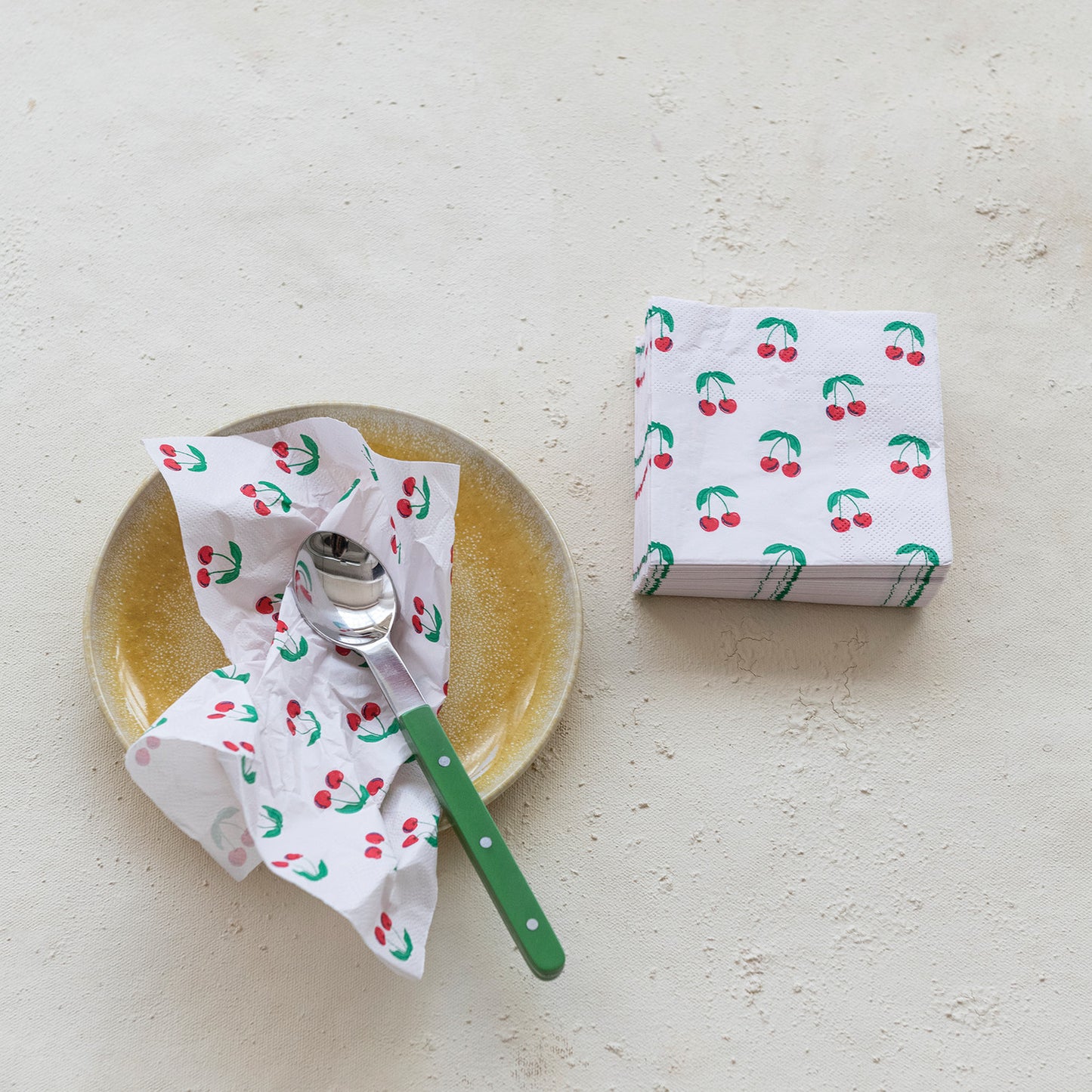Napkin with cherry pattern on a plate with a spoon and a stack of cherry napkins next to it