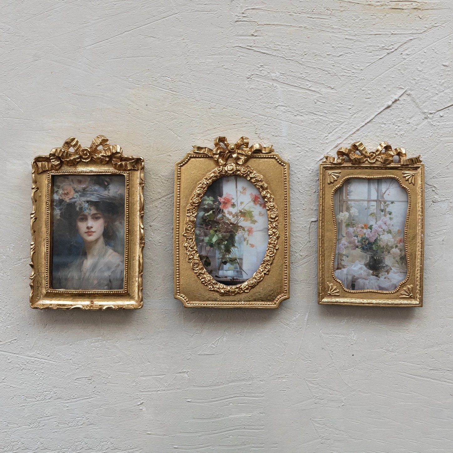 Three ornate gold picture frames with vintage-style artworks on a textured light gray wall.