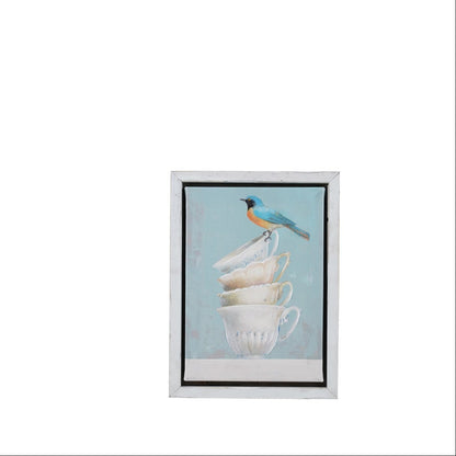 framed art print featuring a small bluebird perched on a stack of white teacups against a light blue background, enclosed in a white frame with a black inner border.