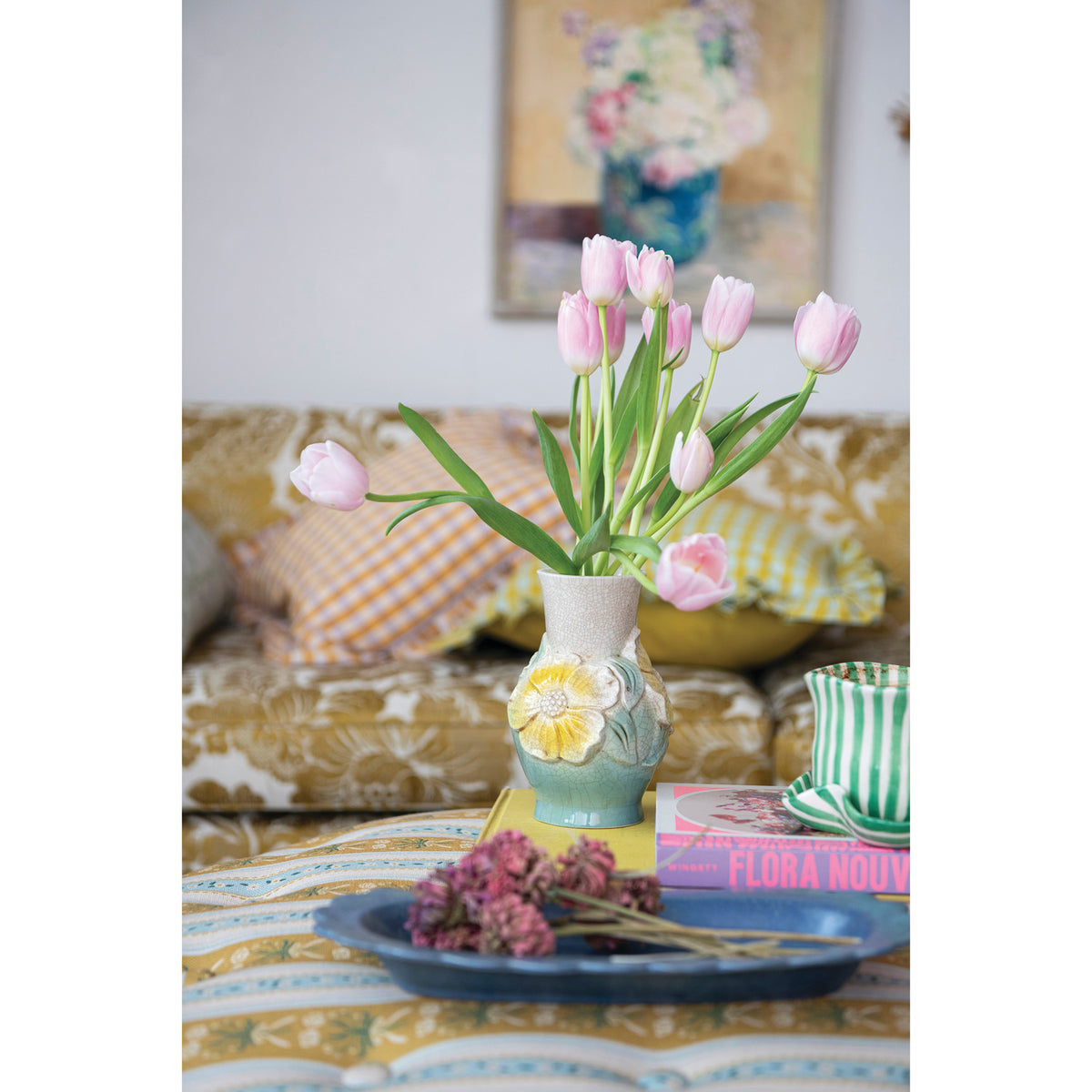 A ceramic vase filled with pink tulips, styled on a patterned tablecloth in a cozy living room setting with pillows and a sofa in the background.