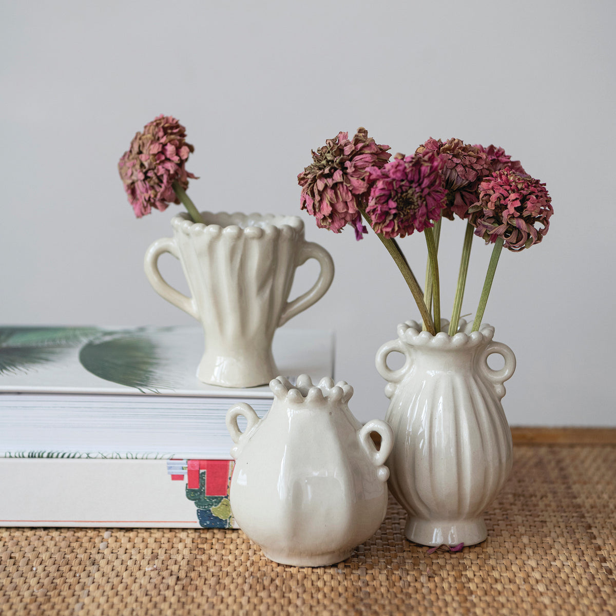 3 assorted small ceramic vases arranged on a table with a stack of books, 2 of the vases have dried flowers in them