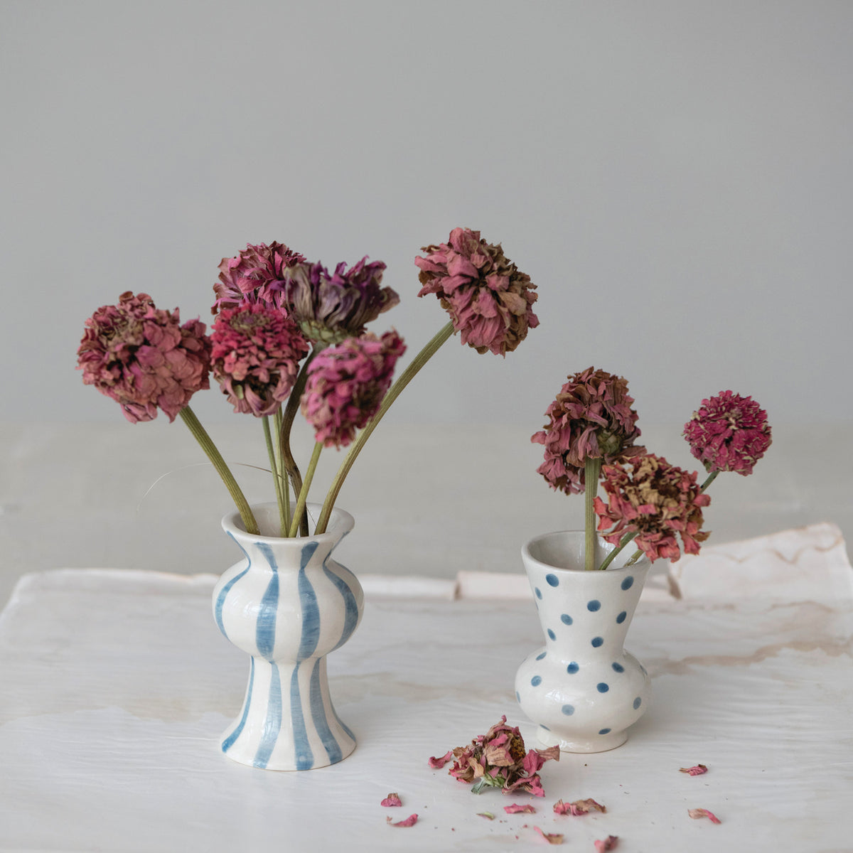 2 small bud vases with blue dots or stripes on them and dried flowers in them set on a white cloth