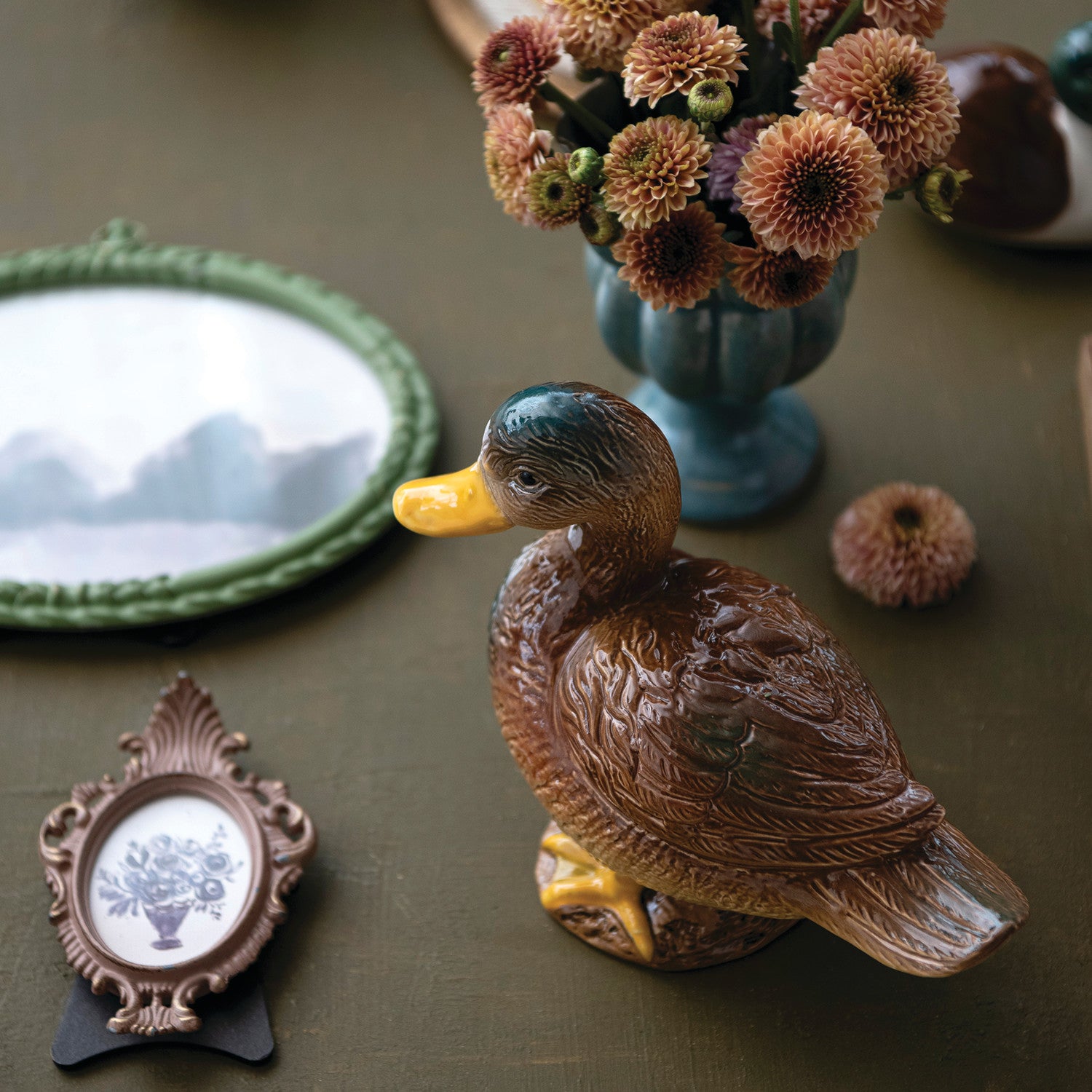 A ceramic duck figurine in brown with a yellow beak and legs, displayed on a dark tabletop alongside a blue flower vase, a green oval mirror, and a small framed picture.