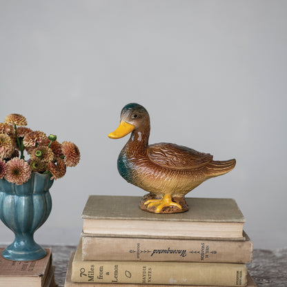 A ceramic duck figurine with a yellow beak and feet, brown body, and teal neck, displayed on top of three vintage books next to a blue vase with dried flowers all shown on a light gray background.