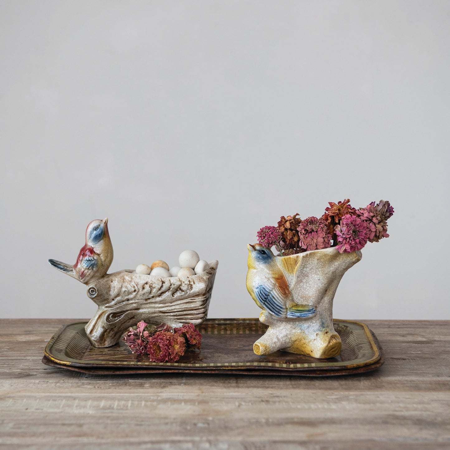 Two ceramic bird-shaped decorative planters on a rectangular tray, one holding white decorative eggs and the other holding pink dried flowers, set on a wooden surface against a neutral gray background.