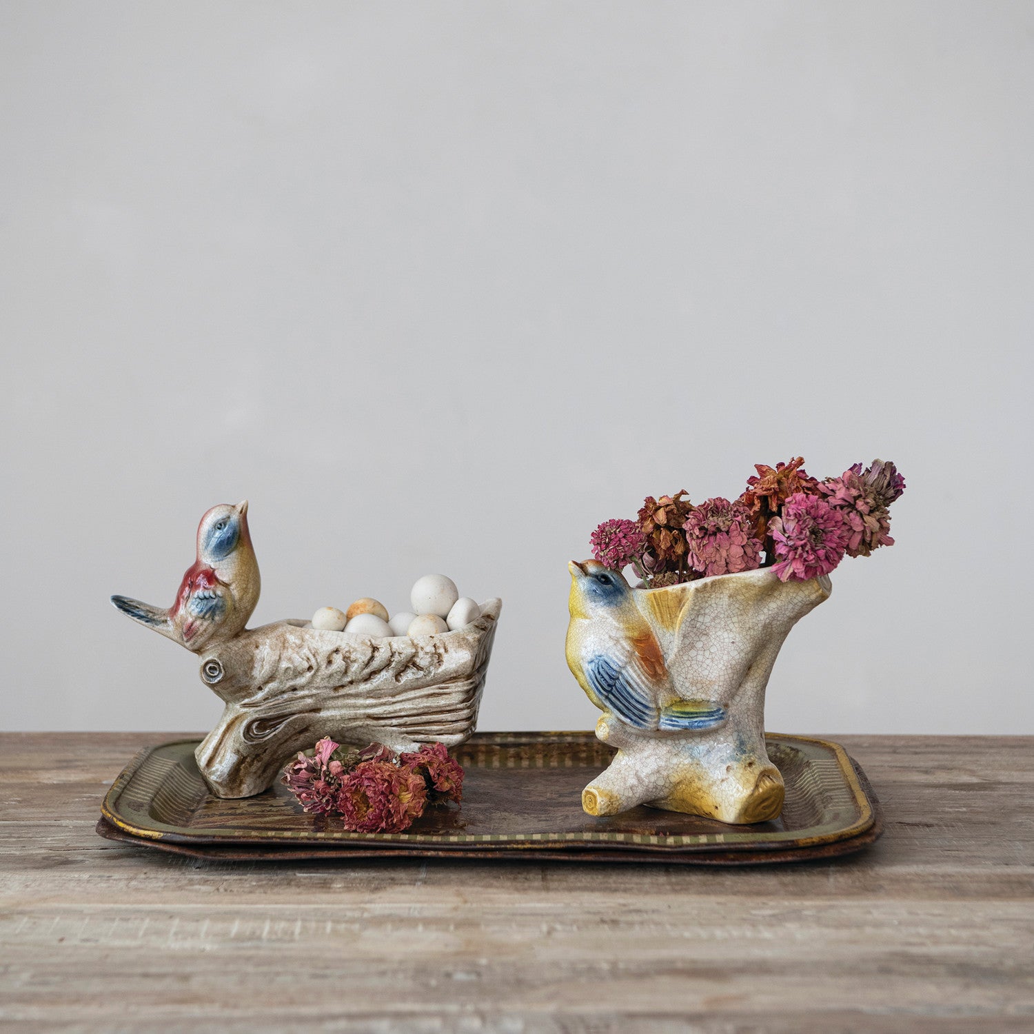 Two ceramic bird-shaped decorative planters on a rectangular tray, one holding white decorative eggs and the other holding pink dried flowers, set on a wooden surface against a neutral gray background.