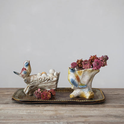 Two ceramic bird-shaped decorative planters on a rectangular tray, one holding white decorative eggs and the other holding pink dried flowers, set on a wooden surface against a neutral gray background.