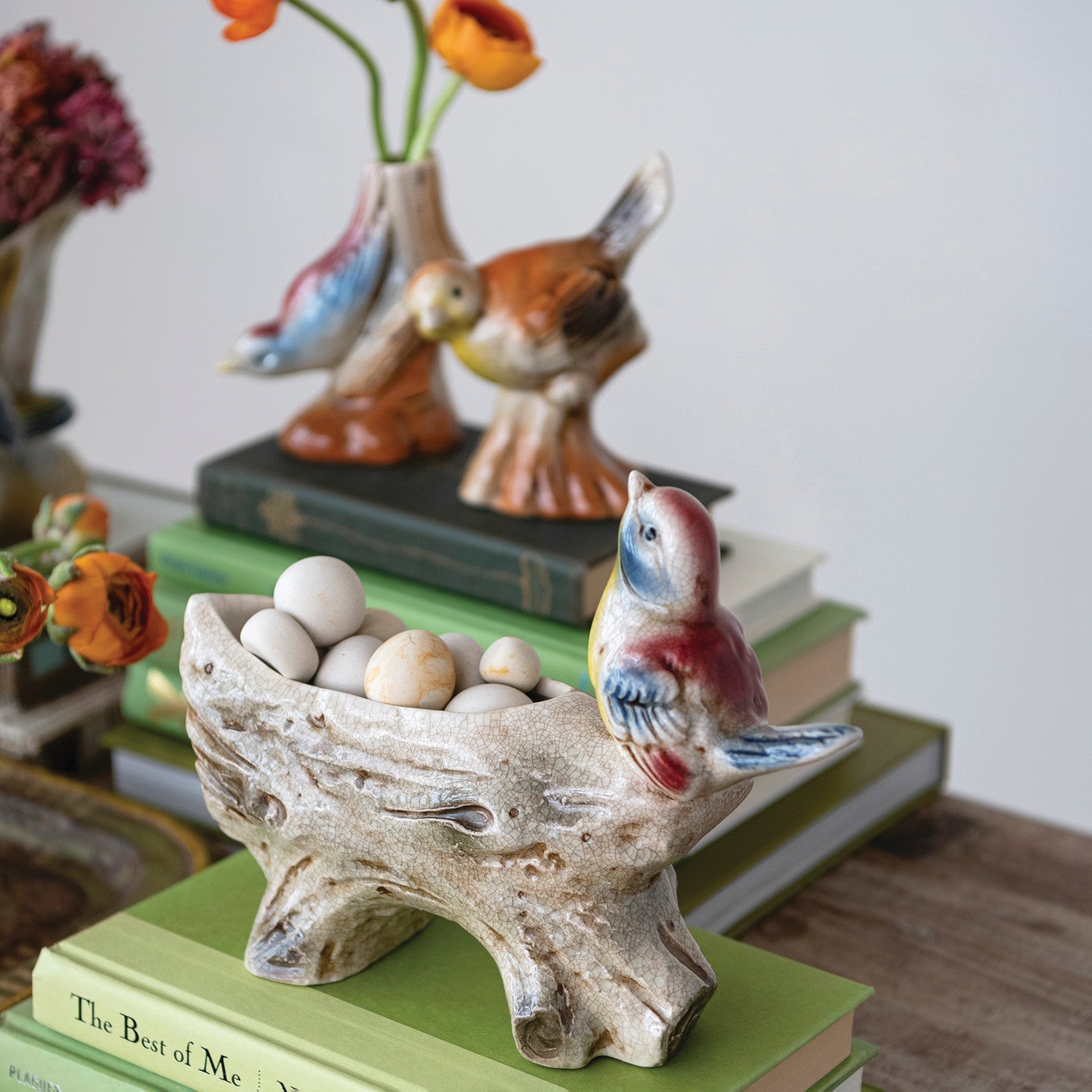 A ceramic decorative bird figurine perched on a tree-branch-shaped tray holding small eggs, styled on top of stacked books with other ceramic birds and flowers in the background.