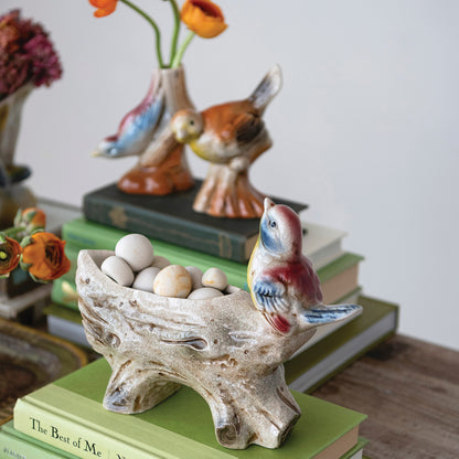 A ceramic decorative bird figurine perched on a tree-branch-shaped tray holding small eggs, styled on top of stacked books with other ceramic birds and flowers in the background.