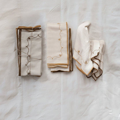 stacks of folded cream-colored linen napkins with brown or mustard embroidered vine details and striped borders, arranged on a textured white fabric background.