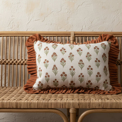 A rectangular decorative throw pillow with a rust brown ruffled border and a cream fabric set on a wicker bench