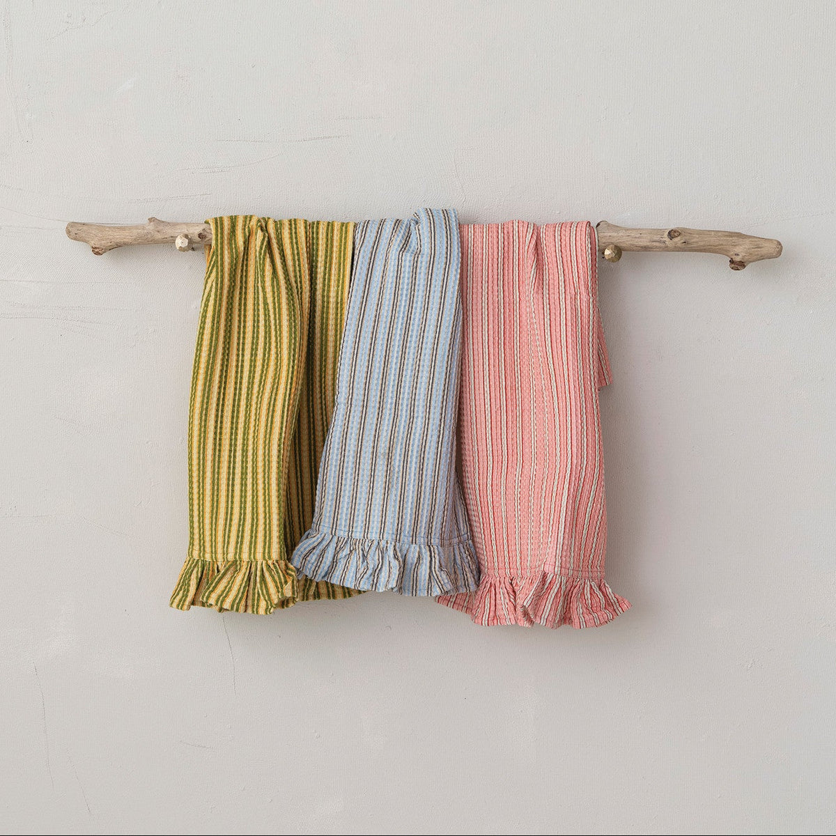 Three striped fabric kitchen towels hanging on a small natural wood branch against a light gray wall. The left towel is olive green and yellow striped, the middle is light blue and white striped, and the right is pink and white striped, all with ruffled hems.