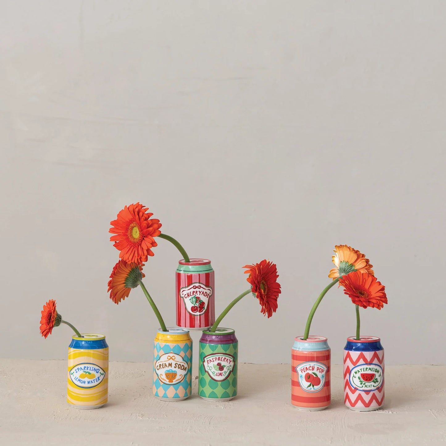 6 miniature ceramic can-shaped flower vases filled with orange and red gerbera daisies, arranged on a light beige surface against a neutral off-white background.