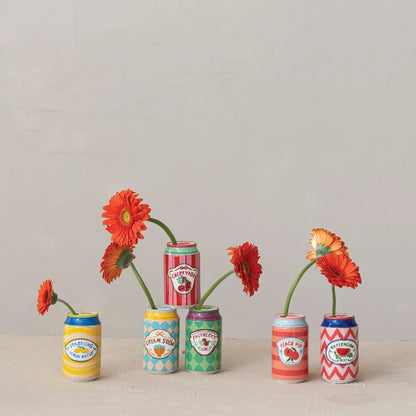 6 miniature ceramic can-shaped flower vases filled with orange and red gerbera daisies, arranged on a light beige surface against a neutral off-white background.