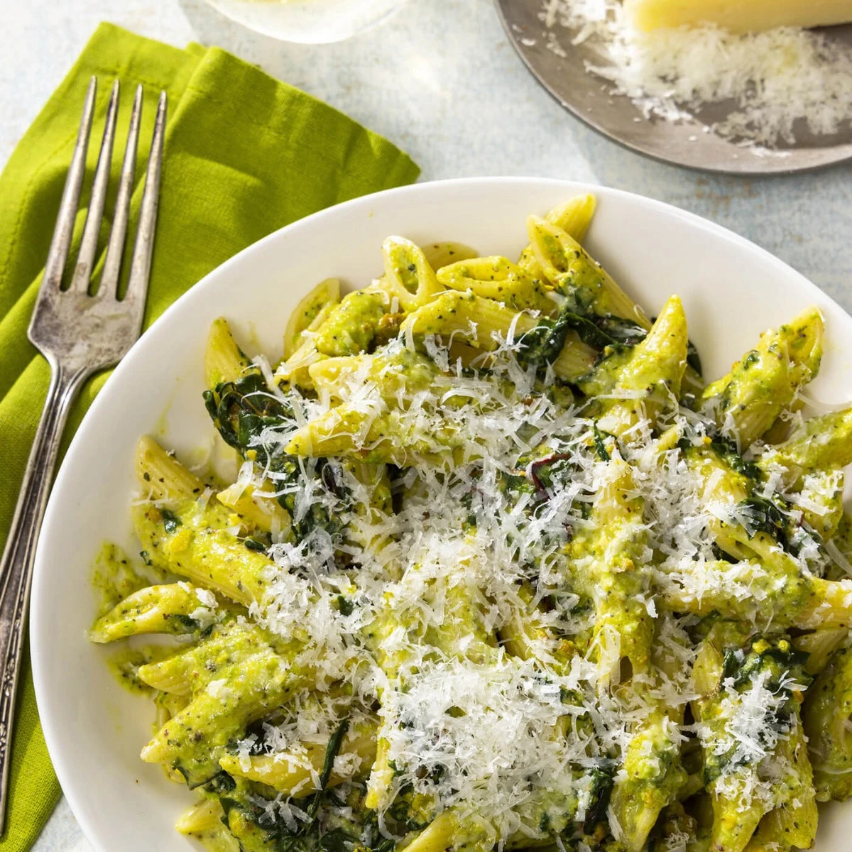 Pasta dish with spinach and parmesan cheese on a white plate with a green napkin and fork.
