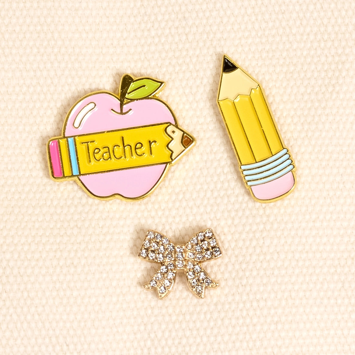 Three pins on a beige background: an apple with 'Teacher' sign, a pencil, and a bow.
