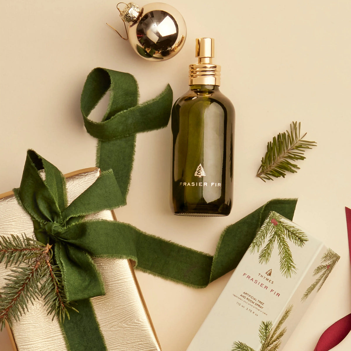 green glass bottle of frasier fir room spray and its box packaging arranged on a beige background with a wrapped gift and a sprig of greenery 