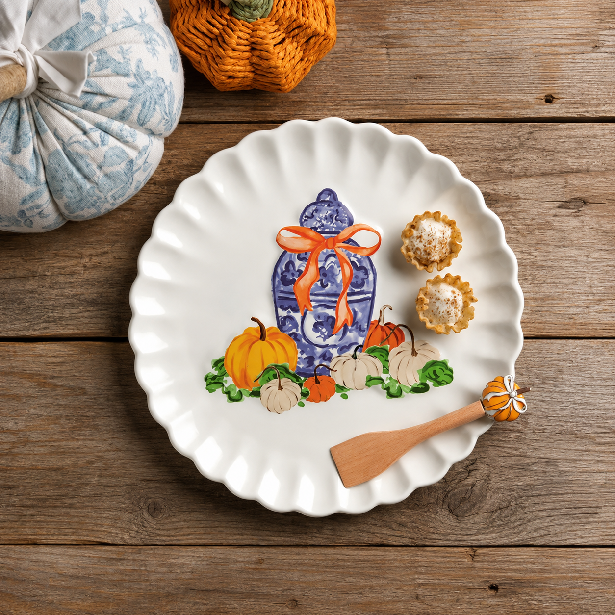 A white scalloped ceramic plate with a blue-and-white floral vase and autumn pumpkin illustration, styled on a rustic wooden surface 