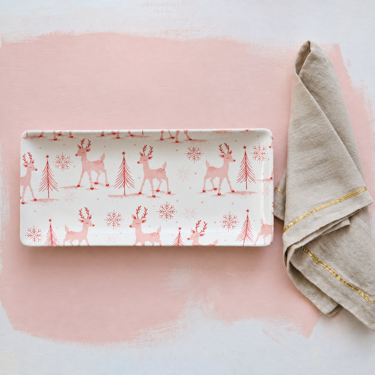 A rectangular ceramic serving tray with a white background and a repeating pattern of pink reindeer, pine trees, and snowflakes, styled on a pink and white background next to a beige napkin with gold trim.