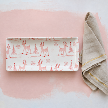 A rectangular ceramic serving tray with a white background and a repeating pattern of pink reindeer, pine trees, and snowflakes, styled on a pink and white background next to a beige napkin with gold trim.