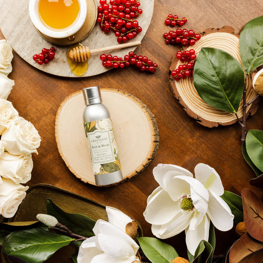 bottle of Birch & Bloom Room Spray laying on a table with greenery, red berries, and honey