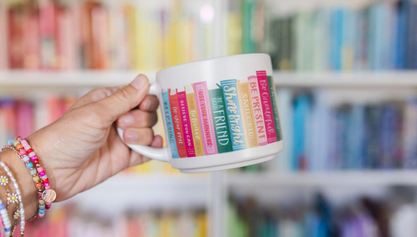 hand holding Affirmation Books Ceramic Mug in front of a book shelf