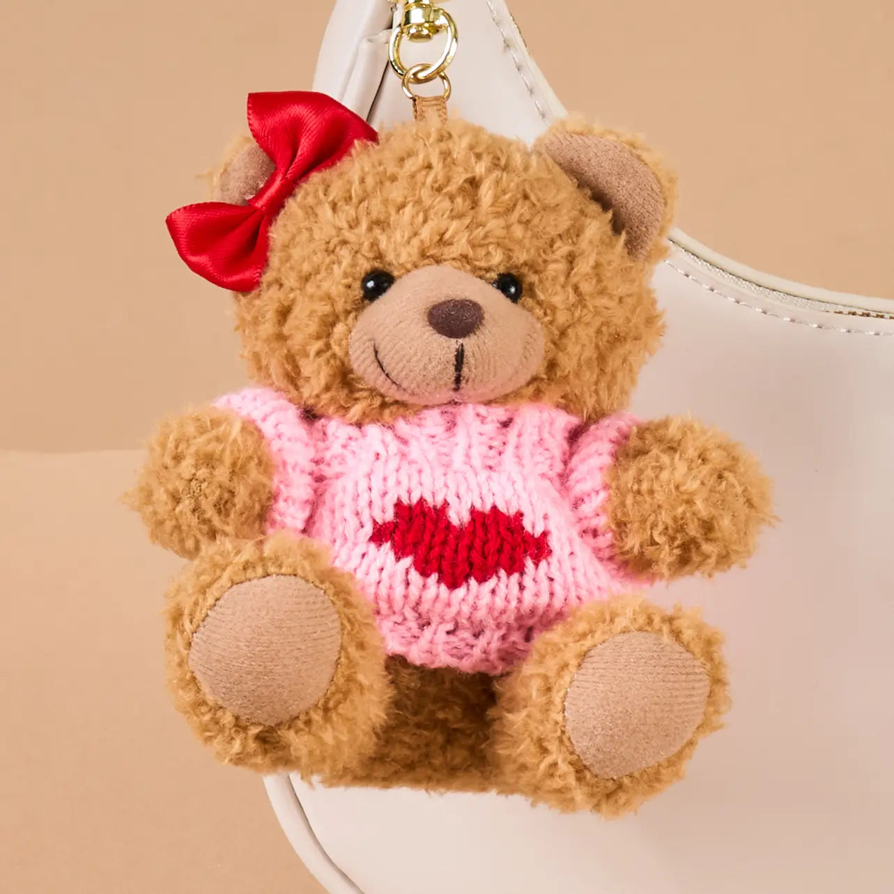 Teddy bear bag charm with a red bow and pink sweater with a heart on clipped on a beige purse