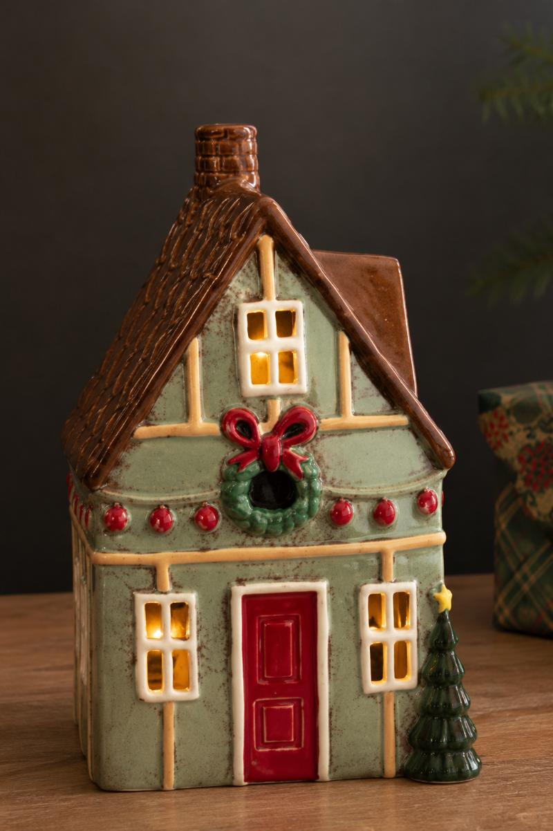 A ceramic Christmas house with a light green facade, brown roof, red door and wreath, and glowing yellow windows, displayed on a wooden surface against a dark background.