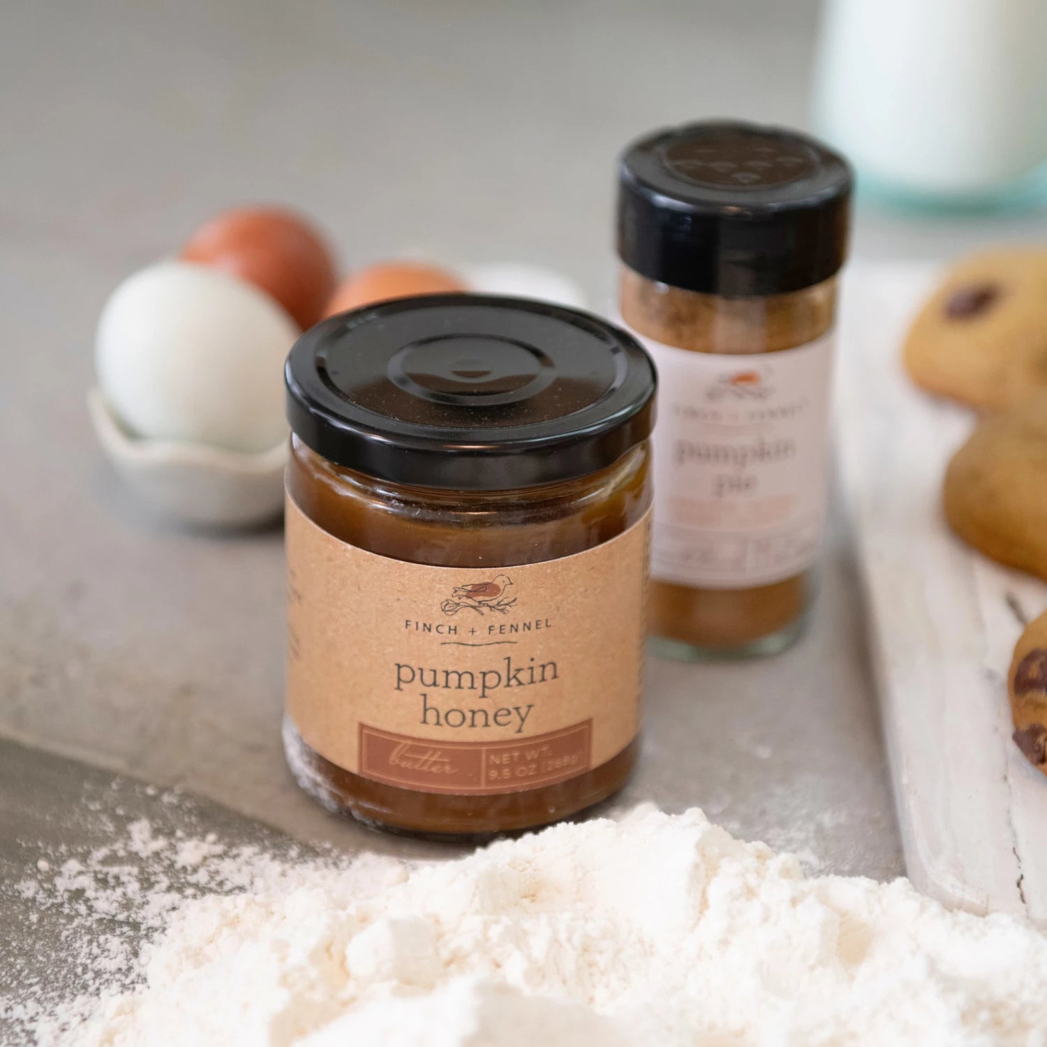 Jar of pumpkin honey with a smaller jar of pumpkin pie spice on a surface with flour and eggs.