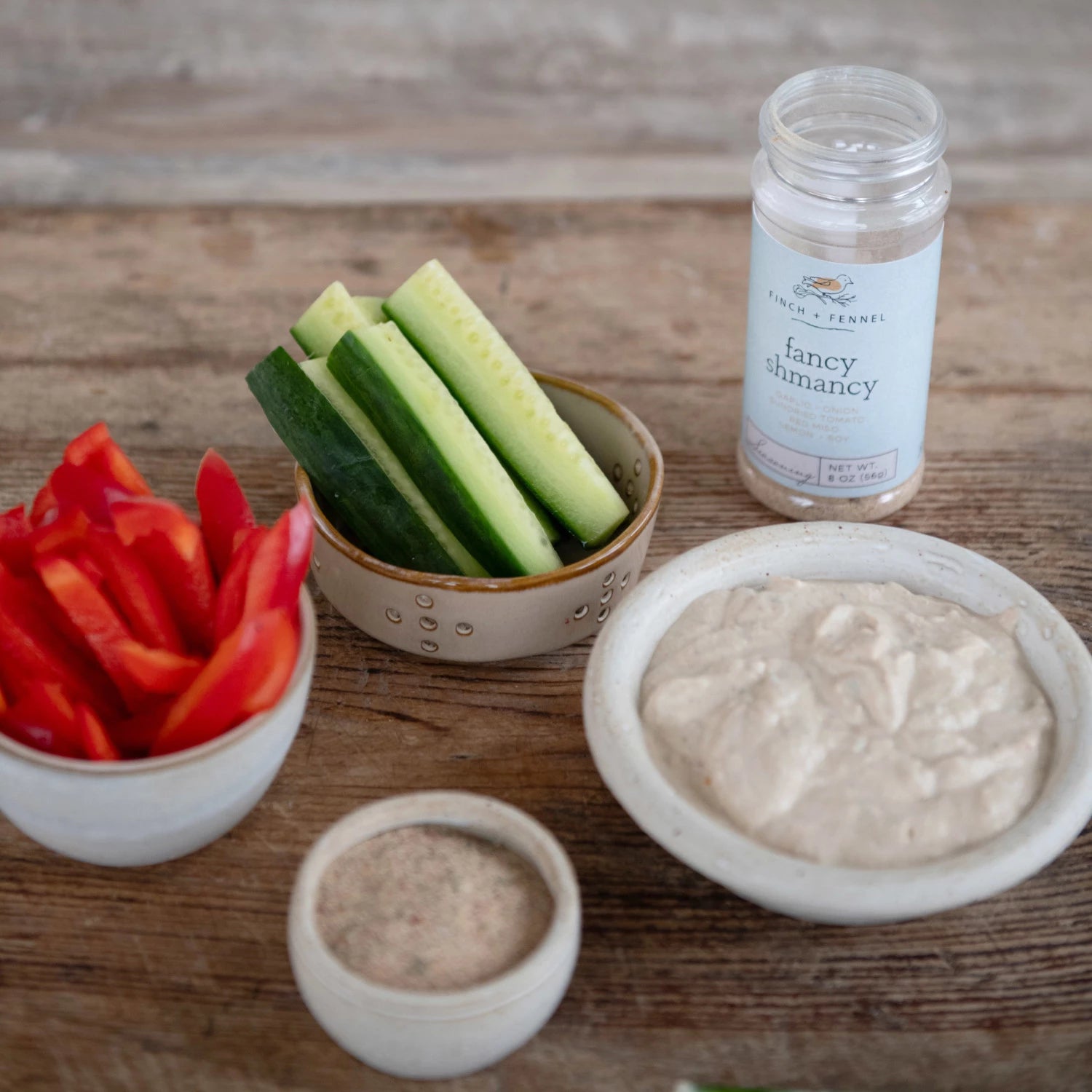 open bottle of Fancy Shmancy Seasoning on a table arranged with bowls of sliced red peppers and cucumber and dip.