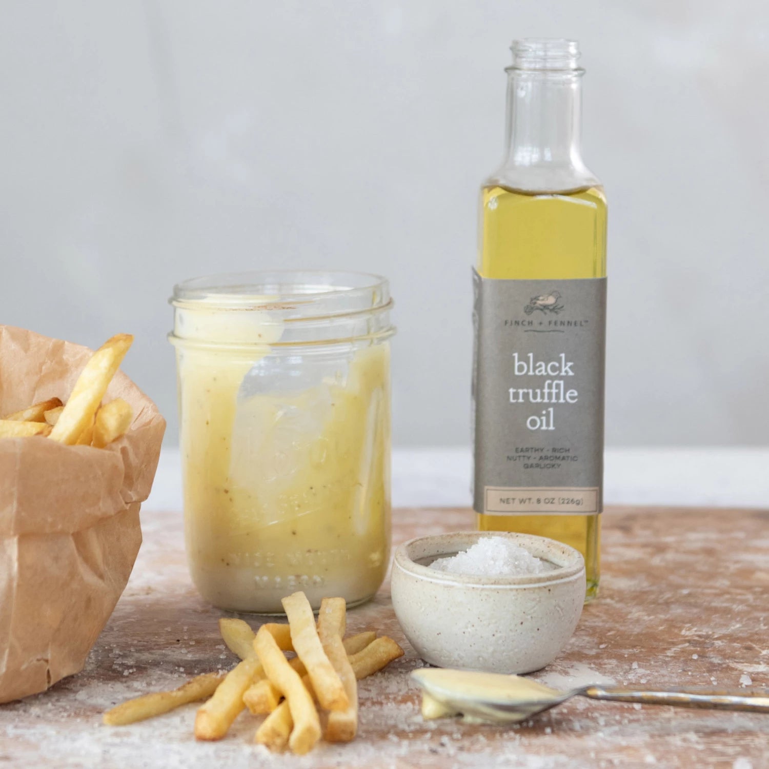 bottle of Black Truffle Oil on a table with a jar of cream, a dish of salt, and french fries acattered ablout.