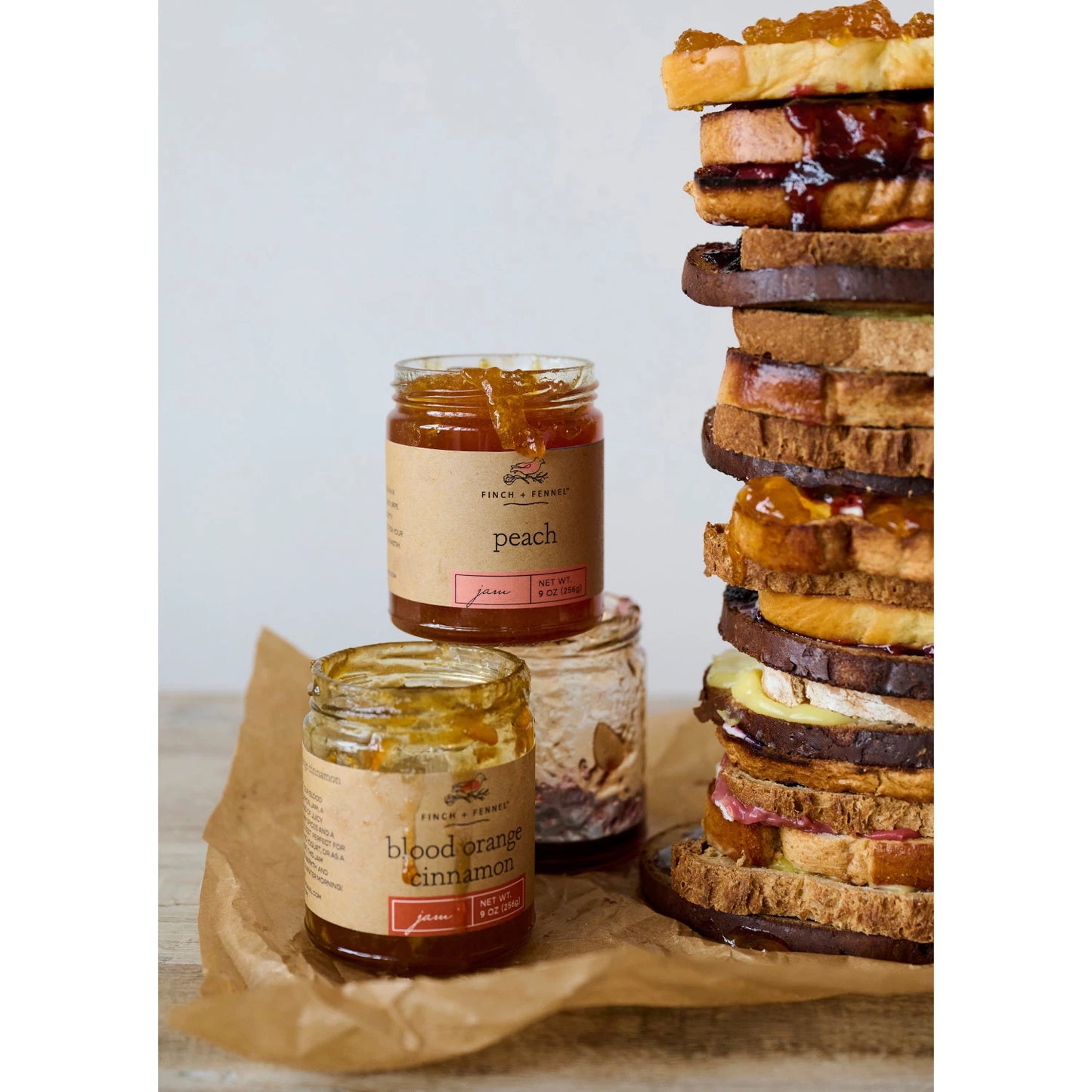Jars of peach and blood orange cinnamon jam with a stack of sandwiches on a wooden surface.