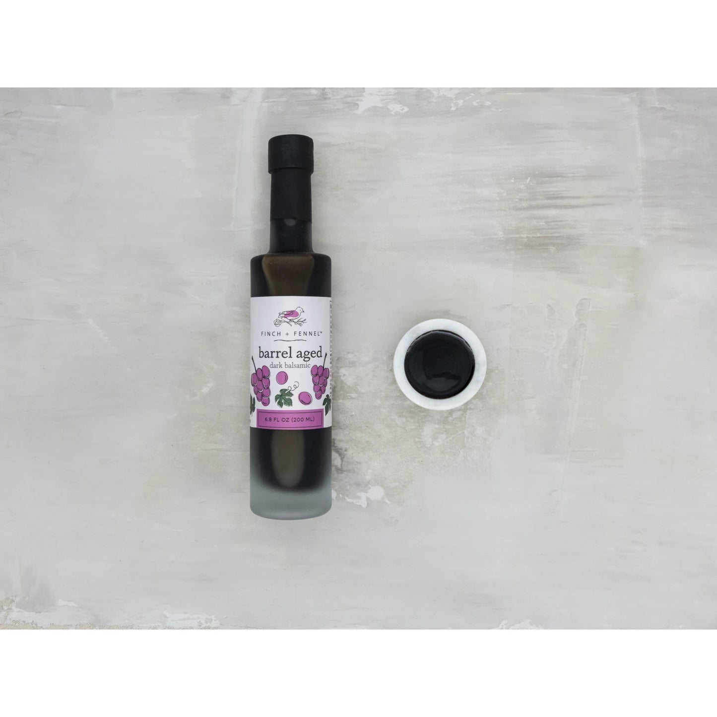 Bottle of barrel-aged balsamic with a small container of it on a light gray background