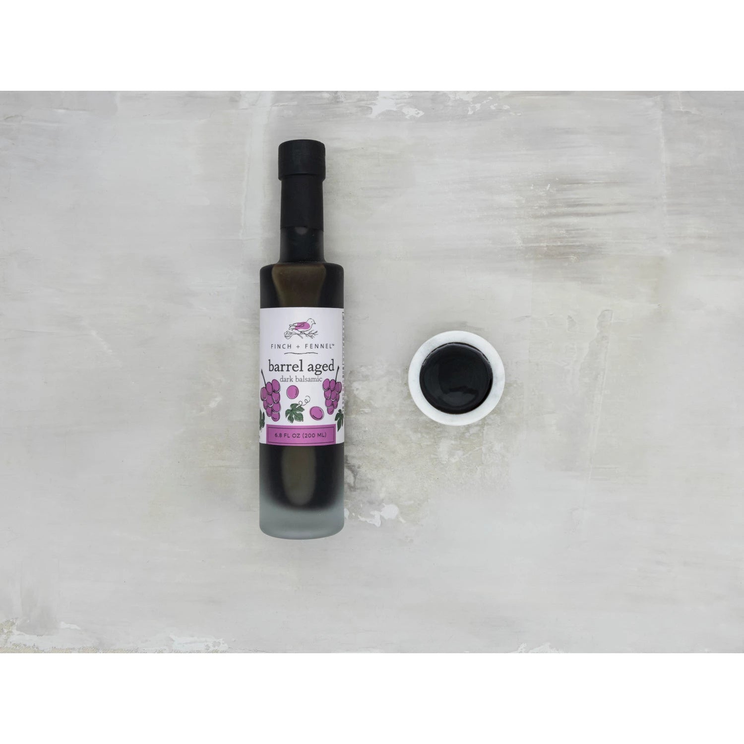 Bottle of barrel-aged balsamic with a small container of it on a light gray background
