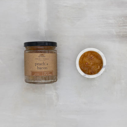 Jar of 'Peach + Bacon' spread next to a small bowl of the spread on a grey marble surface.