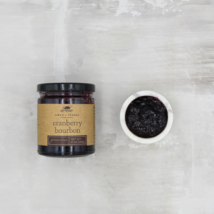 jar of cranberry bourbon preserves set next to a small bowl of cranberry bourbon preserves on a grey surface