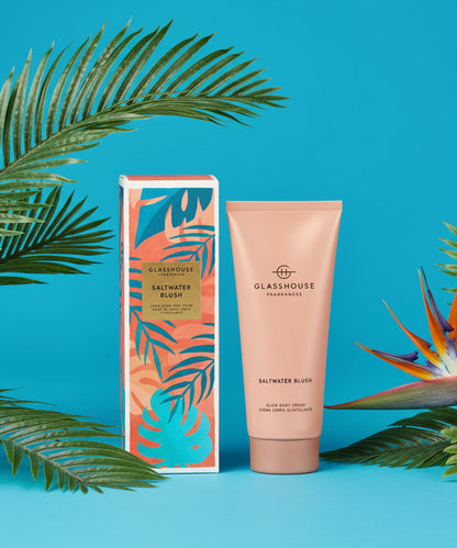 Glasshouse Saltwater Blush body cream and packaging on a blue background arranged with palm leaves and flower.
