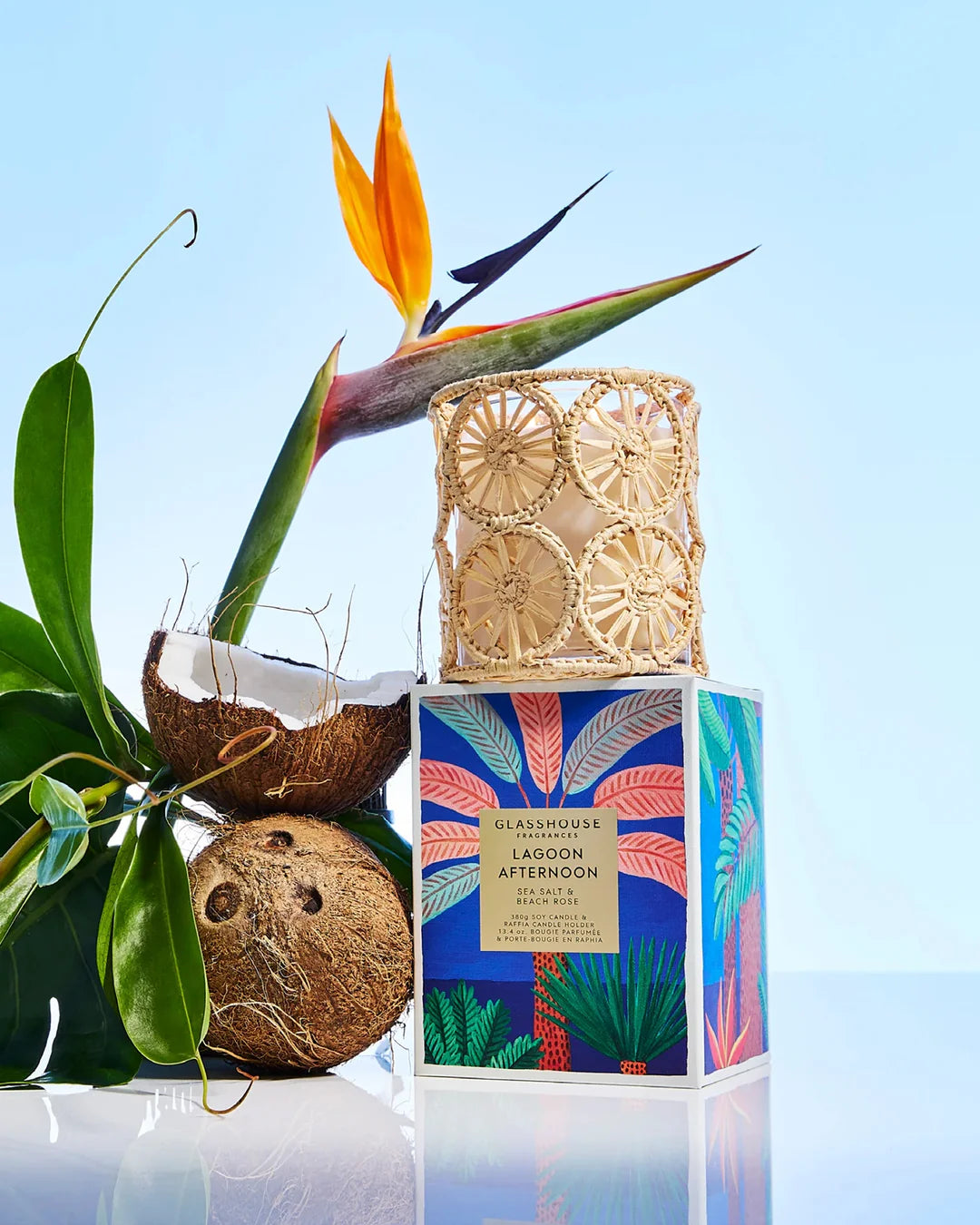 Candle with decorative packaging arranged with a coconut and bird of paradise plant on a reflective surface.