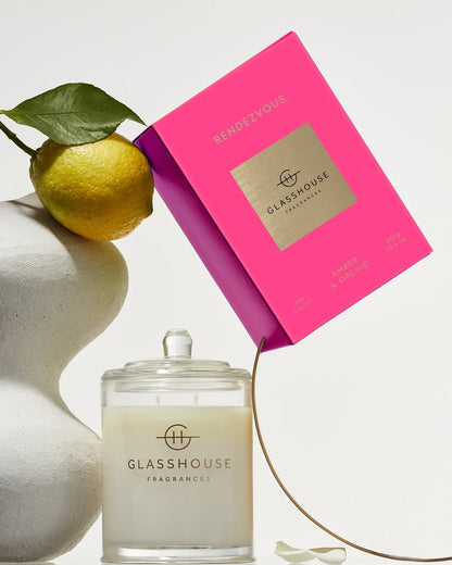 A candle jar with a glass lid and cream wax sits with its bright pink rectangular box. A yellow lemon with a green leaf rests against a white sculptural object on the left.