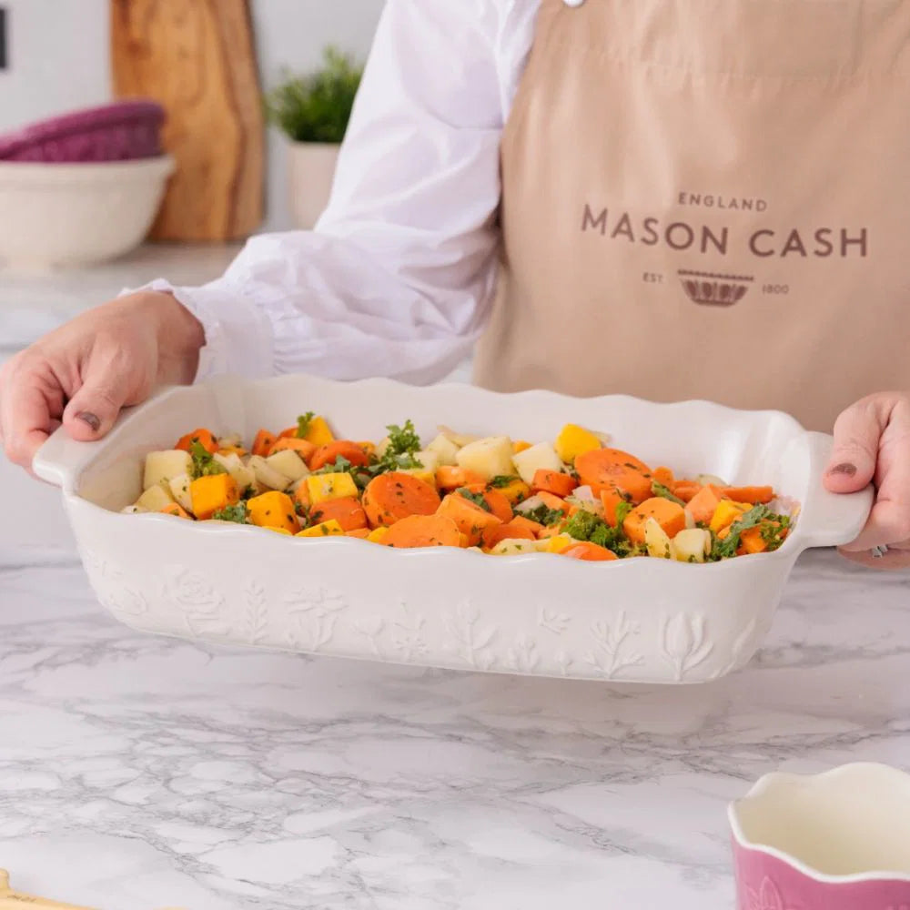 Person preparing a dish with carrots and other vegetables in a white baking dish, wearing a Mason Cash apron.