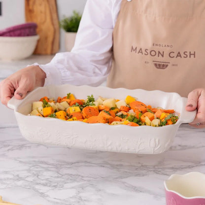 Person preparing a dish with carrots and other vegetables in a white baking dish, wearing a Mason Cash apron.