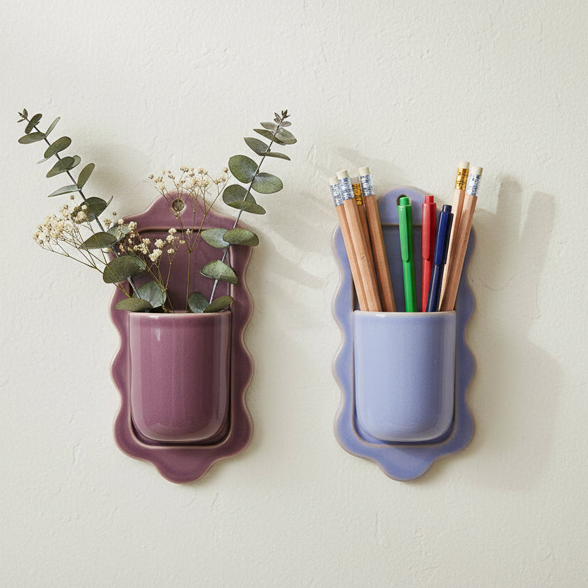 stoneware wall pockets hung on a wall, one purple with greenery in it, one blue with pencils and pens in it 