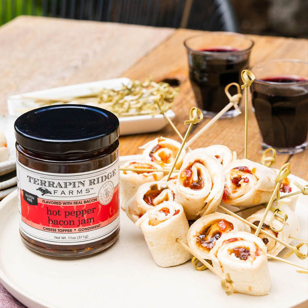 jar of Hot Pepper Bacon Jam with rolled appetizers and glasses of wine in the background.