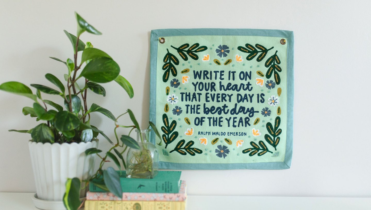light teal "best day" canvas with floral embroidery and darker teal binding hanging on a wall with plants and books next to it
