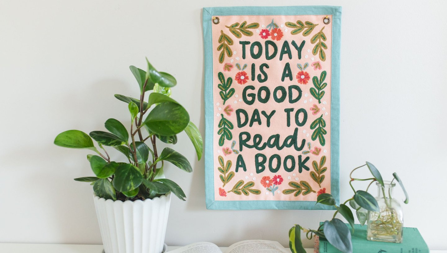 pink "good day" banner with floral embroidery and teal binding hanging on a wall with plants and books set next to it