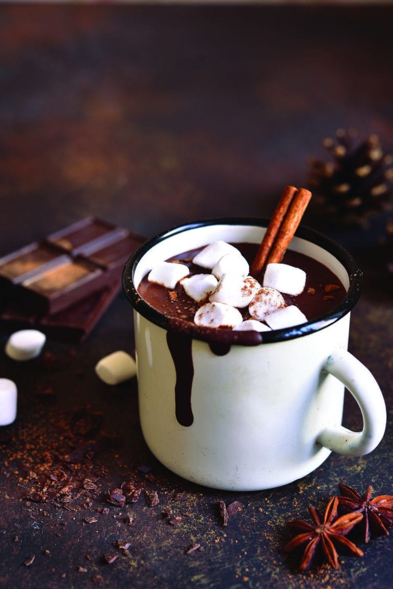 mug of cocoa topped with marshmallows set on a table surrounded by spices and chocolate bars.