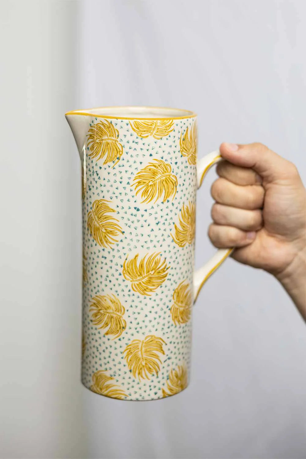 Hand holding a ceramic pitcher with yellow leaf pattern on a plain background