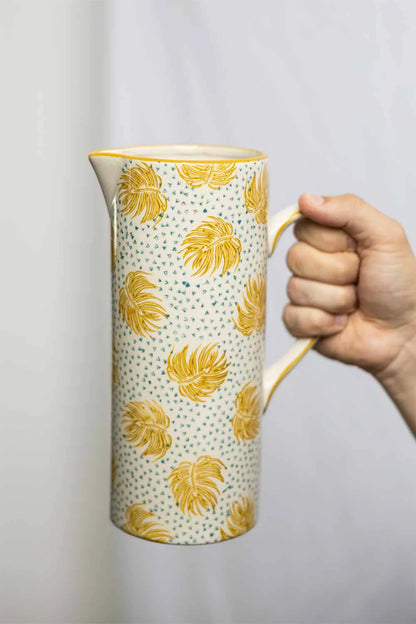 Hand holding a ceramic pitcher with yellow leaf pattern on a plain background