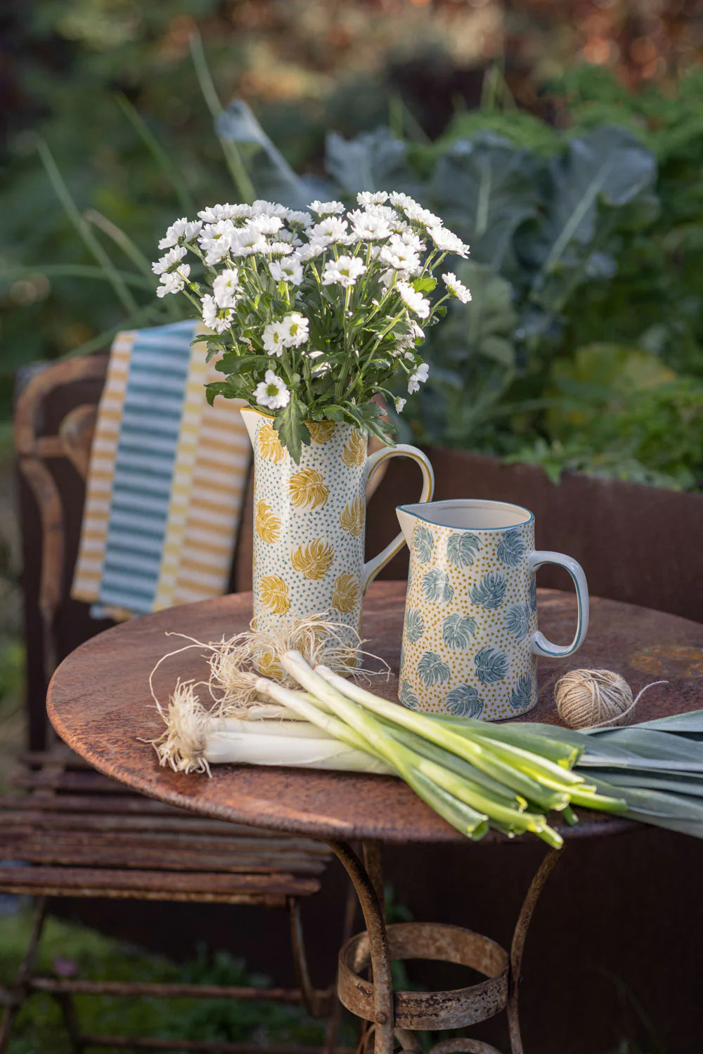 pitcher filled with flowers set on a wooden table outdoors with another pitcher and veggies
